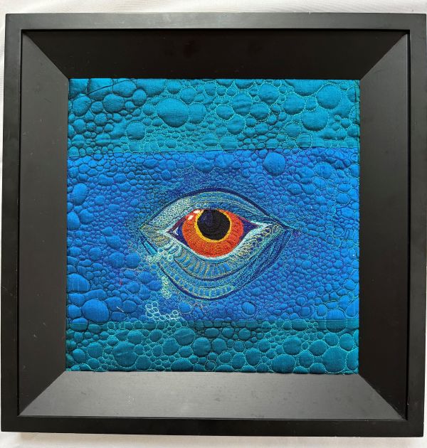 Robin DeMuth Schofield - Lizard Views (quilt in a black frame. it is all blue with a bright eye in the center and a pebbly quilting pattern like lizard skin)