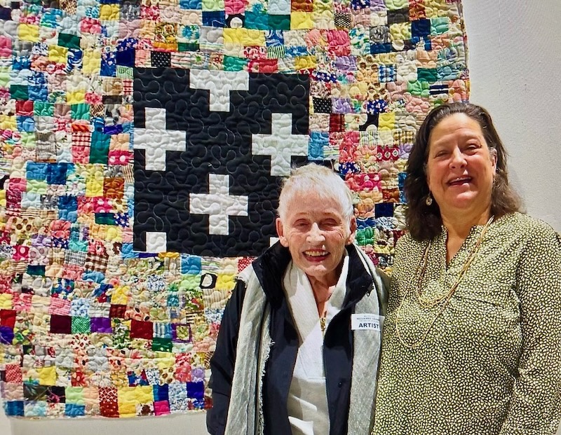 Sandy Teepen - Raw Edge Collage (Sandy Teepen and friend stand in front of a quilt made of a central block of white crosses on black surrounded by small colorful squares.)