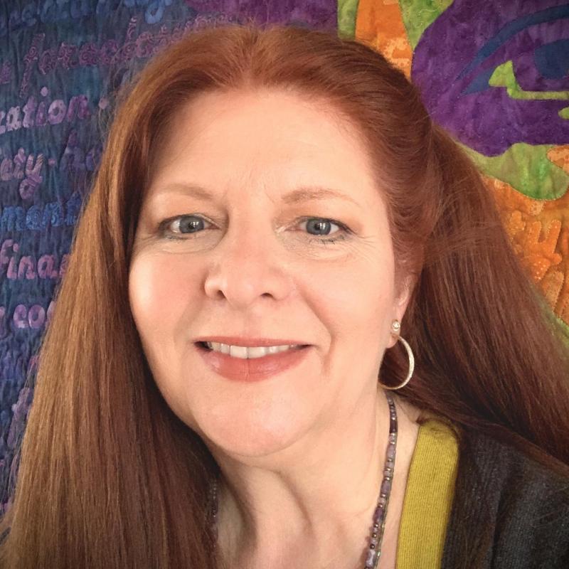 Michelle Jackson is looking at the camera and smiling. She has red hair and a green shirt. There is artwork behind her that is purple and green.
