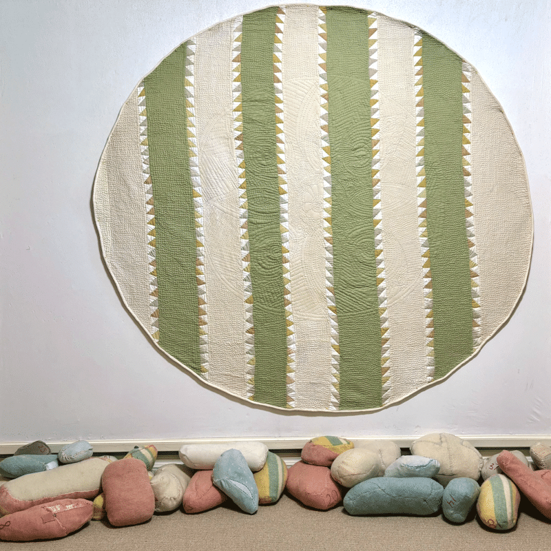 Judy Martin - The Beauty of the Green Earth with Mothering Bundles (a green and white striped circular quilt hangs above quilted rock-like bundles)