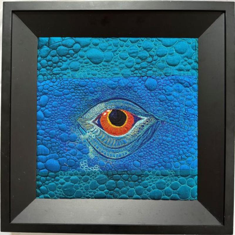 Robin DeMuth Schofield - Lizard Views (quilt in a black frame. it is all blue with a bright eye in the center and a pebbly quilting pattern like lizard skin)