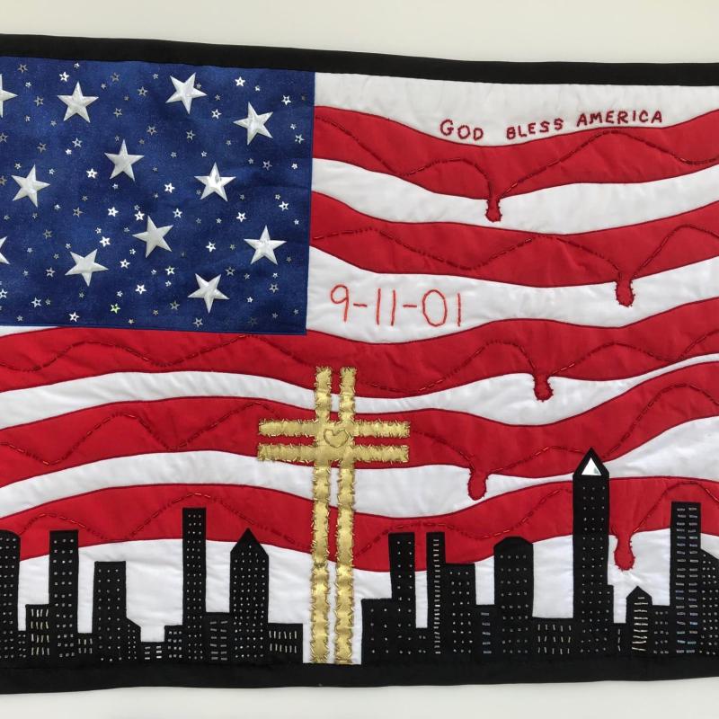 Susan Hoppenworth - 25 Years - 9/11 Remembered (the NYC skyline over an edited American flag that reads "God Bless America" and "9-11-01". A golden cross with heart is in the center and the red stripes of the flag are dripping)