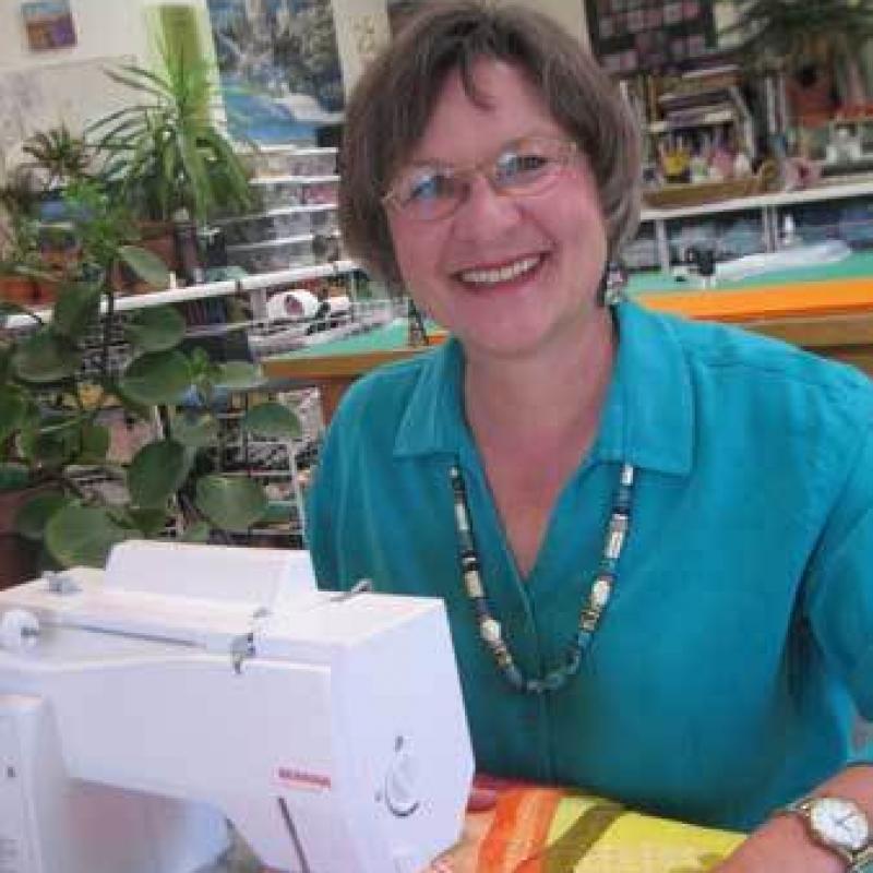 Katie Pasquini Masopust sitting at her sewing machine. She is smiling and wearing a teal shirt. There is a colorful quilt under the needle of the machine.